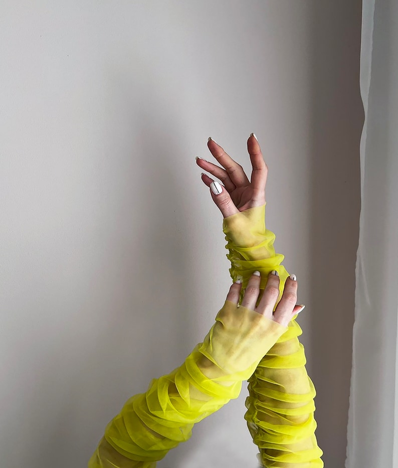 May include: A pair of bright yellow mesh long sleeves with ruched detailing. The sleeves are worn on a person's arms, with the hands visible.