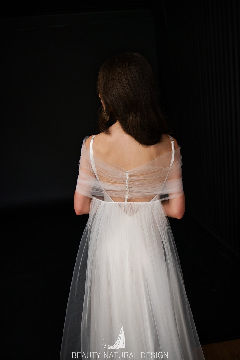 Puede incluir: Un vestido de novia de tul blanco con la espalda baja y una superposici&oacute;n transparente. El vestido tiene una fila de botones en la espalda y una falda fluida.