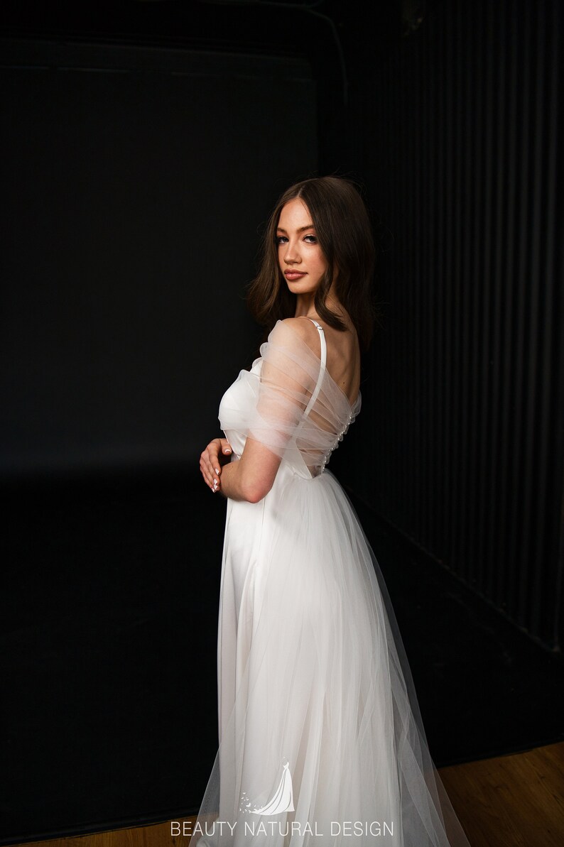 Puede incluir: Un vestido de novia de tul blanco con un corpi&ntilde;o de sat&eacute;n y una superposici&oacute;n transparente. El vestido tiene un escote de coraz&oacute;n y un corpi&ntilde;o ajustado. La falda es completa y fluida.