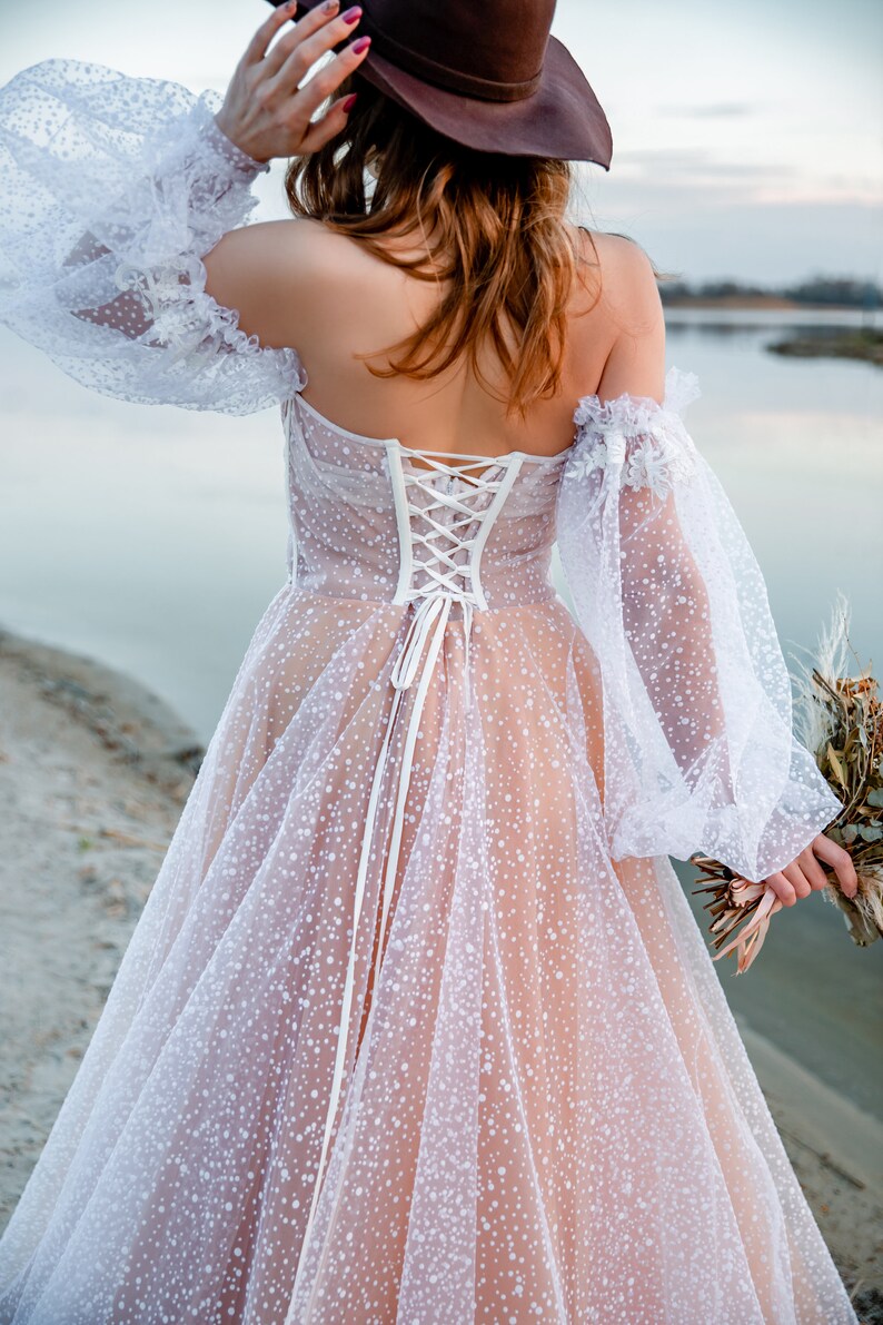 A white tulle wedding dress with a lace bodice and a fitted corset back. The dress has a scattered dot pattern and is worn with a brown cowboy hat.