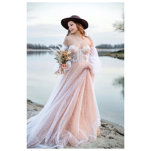Boho bride in a lace corset gown and brown hat holding a bouquet, standing by a river at sunset. Rustic wedding inspiration and alternative bridal style.