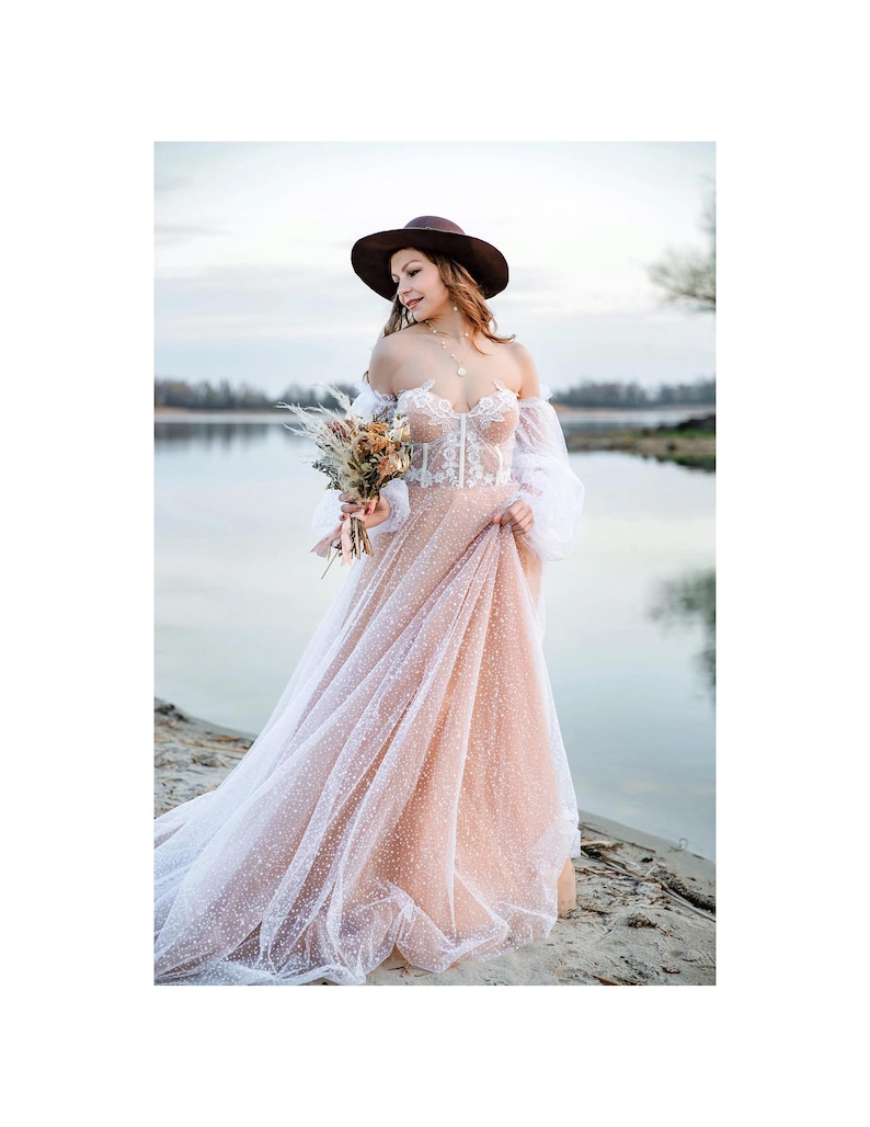 Boho bride in a lace corset gown and brown hat holding a bouquet, standing by a river at sunset. Rustic wedding inspiration and alternative bridal style.
