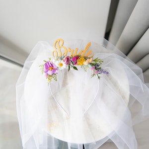 May include: A white bridal veil with a gold "Bride" headband adorned with a floral crown of pink, purple, yellow, and white flowers.