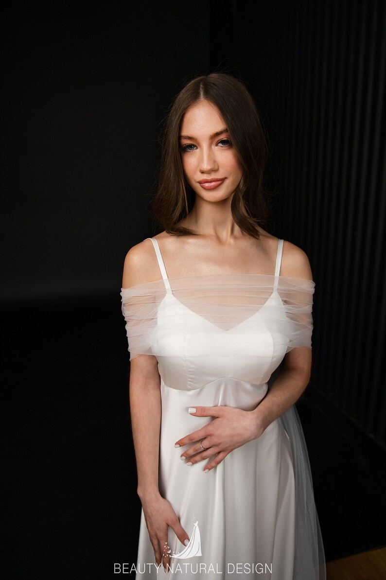 Puede incluir: Un vestido de novia blanco de hombros descubiertos con una capa de tul y un corpi&ntilde;o de sat&eacute;n. El vestido tiene un dise&ntilde;o simple y elegante, perfecto para una novia que quiere un look cl&aacute;sico.