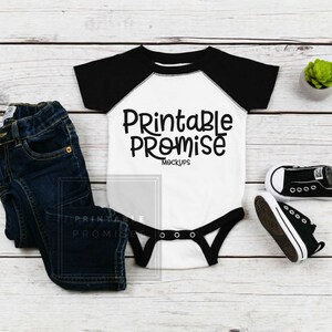 May include: A white baby onesie with black raglan sleeves and the text "Printable Promise Mockups" on the front. The onesie is laying on a white wooden surface with a pair of black baby shoes and a small potted plant.