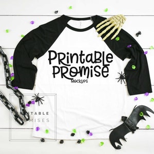 May include: A white and black raglan sleeve t-shirt with the text "Printable Promise Mockups" in black. The shirt is decorated with Halloween-themed accessories, including a skeleton hand, spiders, and a bat.