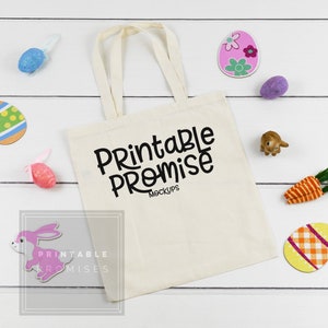 Op de afbeelding: Een witte tote bag met zwarte tekst die "Printable Promise Mockups" zegt. De tas is omringd door paaseieren, een roze konijn en een wortel.