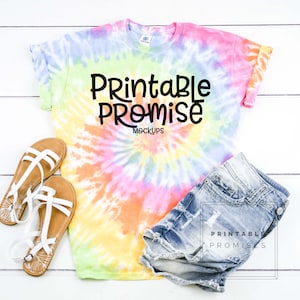 May include: A tie-dye t-shirt with the text "Printable Promise Mockups" is displayed with denim shorts and white sandals. The shirt has a spiral pattern in shades of pink, yellow, green, and blue. The shorts are light blue denim.