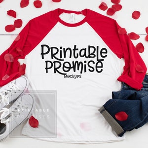 May include: A white and red baseball tee with the text "Printable Promise Mockups" in black. The shirt is paired with white sneakers, blue jeans, and red rose petals scattered on a white wooden surface.
