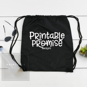 May include: Black drawstring bag with white text that reads "Printable Promise Mockups". The bag is on a white wooden surface with a clear water bottle, sunglasses, and two small potted plants.