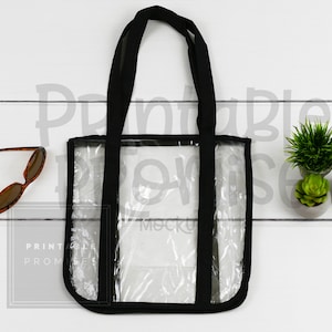 Puede incluir: Una bolsa de mano transparente con correas negras y un ribete negro. La bolsa está hecha de plástico transparente y tiene un cierre de cremallera negro. La bolsa está sobre una superficie de madera blanca.