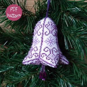 May include: Purple and white cross-stitch bell ornament with a snowflake pattern. The bell is hanging from a green pine branch. PDF pattern included.