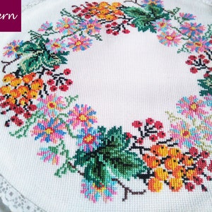 Peut inclure: Une nappe ronde blanche avec une bordure en dentelle crochetée. Le centre de la nappe présente une couronne florale colorée avec des baies rouges, des fleurs jaunes et des feuilles vertes.