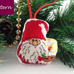 May include: A red and white cross-stitch ornament of a gnome holding a candle. The gnome is wearing a red hat and has a white beard. The ornament is hanging from a gold chain.