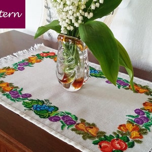 May include: White table runner with a fringed border featuring a colourful embroidered design of fruit and leaves. The runner is on a wooden table with a vase of white flowers.