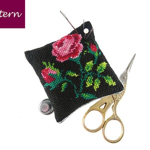May include: A black pincushion with a cross-stitch design of pink and red roses and green leaves. The pincushion has a small metal thimble and a pair of gold scissors.
