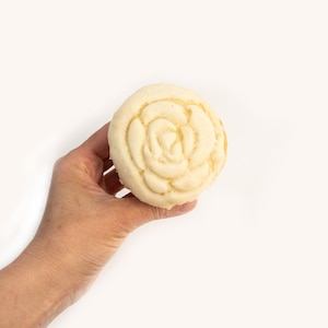 Op de afbeelding: Een rond, wit gebakken product met een roosvormig ontwerp aan de bovenkant. Het gebakken product wordt in een hand gehouden.
