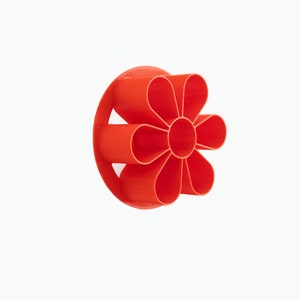 May include: A red plastic flower-shaped cookie cutter. The cutter has eight petals and a circular center.