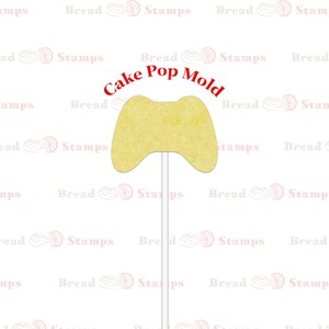 Puede incluir: Un molde amarillo para cake pops con forma de mando de videojuegos. Las palabras "Cake Pop Mold" están impresas encima del mando. El molde está unido a un fino palito blanco, listo para sumergir y decorar.