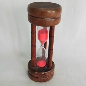 Può includere: Una clessidra in legno marrone scuro. La sezione centrale è in vetro trasparente, riempita di sabbia rossa. La clessidra ha una parte superiore e inferiore cilindrica, unite da quattro supporti verticali in legno. Oggetto decorativo.