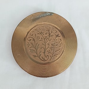 May include: A round, antique brass compact with a floral design in the center. The compact has a ridged outer edge and a slightly worn appearance, suggesting age and use. The central design features an embossed pattern of leaves and flowers.