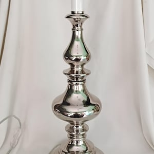 May include: A polished silver table lamp with an ornate design. The lamp has a bulbous base, a central column with rounded shapes, and a white cylindrical light fixture at the top. A power cord with an inline switch extends from the base.