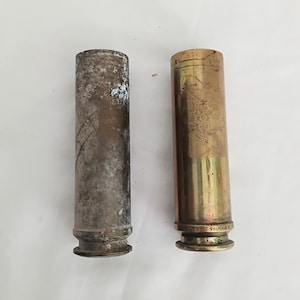 May include: Two vintage brass and metal shell casings. One is tarnished with a silver and brown patina, the other is a golden brass colour. The base of the brass casing is stamped with text.