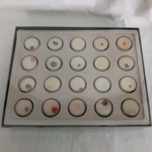 Puede incluir: Una vitrina rectangular con marco negro e interior gris. La caja contiene veinti&uacute;n peque&ntilde;os recipientes redondos y transparentes, cada uno con una muestra diferente de piedra preciosa o mineral. Las muestras var&iacute;an en color y textura.