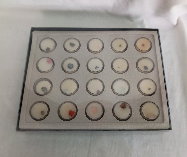 Puede incluir: Una vitrina rectangular con marco negro e interior gris. La caja contiene veinti&uacute;n peque&ntilde;os recipientes redondos y transparentes, cada uno con una muestra diferente de piedra preciosa o mineral. Las muestras var&iacute;an en color y textura.