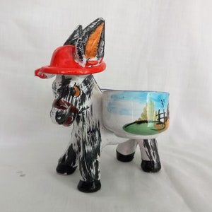 May include: A ceramic donkey salt and pepper shaker set. The donkey is painted white, black, and orange, and is wearing a red hat. The salt and pepper shakers are attached to the donkey's back and are painted with a blue sky, green grass, and a fence.