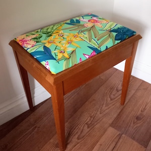May include: A wooden stool with a floral patterned cushion. The cushion features a tropical design with green leaves, yellow flowers, and pink accents. The stool has a light brown finish and four legs.