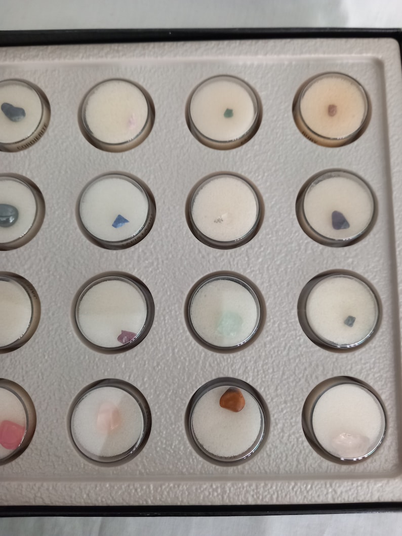 Puede incluir: Una colecci&oacute;n de peque&ntilde;as piedras preciosas de colores exhibidas en una bandeja gris con compartimentos circulares transparentes. Cada gema est&aacute; colocada sobre una base de espuma blanca. Las piedras preciosas var&iacute;an en color, incluyendo azul, rosa, verde y marr&oacute;n.