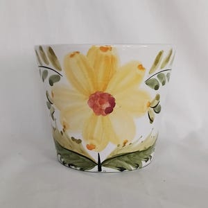 May include: White ceramic flower pot featuring a large yellow daisy design. The flower has a red centre and is surrounded by green leaves and stems. The pot is approximately 15 cm tall.
