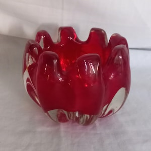 Può includere: Un posacenere in vetro rosso vibrante con un design unico a forma di petalo. Il posacenere presenta una base trasparente e arrotondata e una finitura lucida. L'interno è rosso intenso, con bordi chiari e traslucidi. Un oggetto decorativo.
