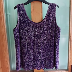 May include: A sleeveless purple top with a scoop neckline, decorated with silver beads in a leaf pattern. The top is suspended on a black hanger. The hemline has a scalloped edge. The top is hanging on a wooden door.
