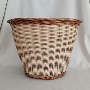 May include: A round, tapered basket woven from light beige wicker material. The basket features a thick, dark brown braided rim at the top and a matching dark brown braided base, creating a distinct contrast with the lighter body of the container.