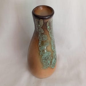 May include: A ceramic vase with a brown rim and a light brown body. The vase has a textured, turquoise and brown pattern on the side. The vase is a decorative item.