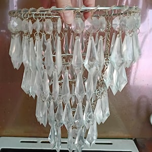 May include: A tiered chandelier shade featuring multiple rows of clear, faceted, diamond-shaped glass crystals suspended from a circular silver-toned metal frame. The crystals are attached with small metal rings, creating a cascading effect.