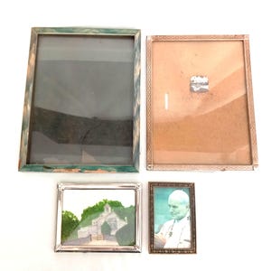 Puede incluir: Cuatro marcos de fotos, dos con un acabado de madera marrón claro, uno con un acabado verde y azul y uno con un acabado plateado. Los marcos son de diferentes tamaños y formas. Los marcos están vacíos.