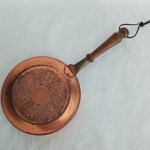 May include: A round copper pan featuring an intricate embossed floral pattern on its base. The pan includes a long, tapered wooden handle with a brass-coloured metal connector and a thin black cord loop attached to the end for convenient hanging.