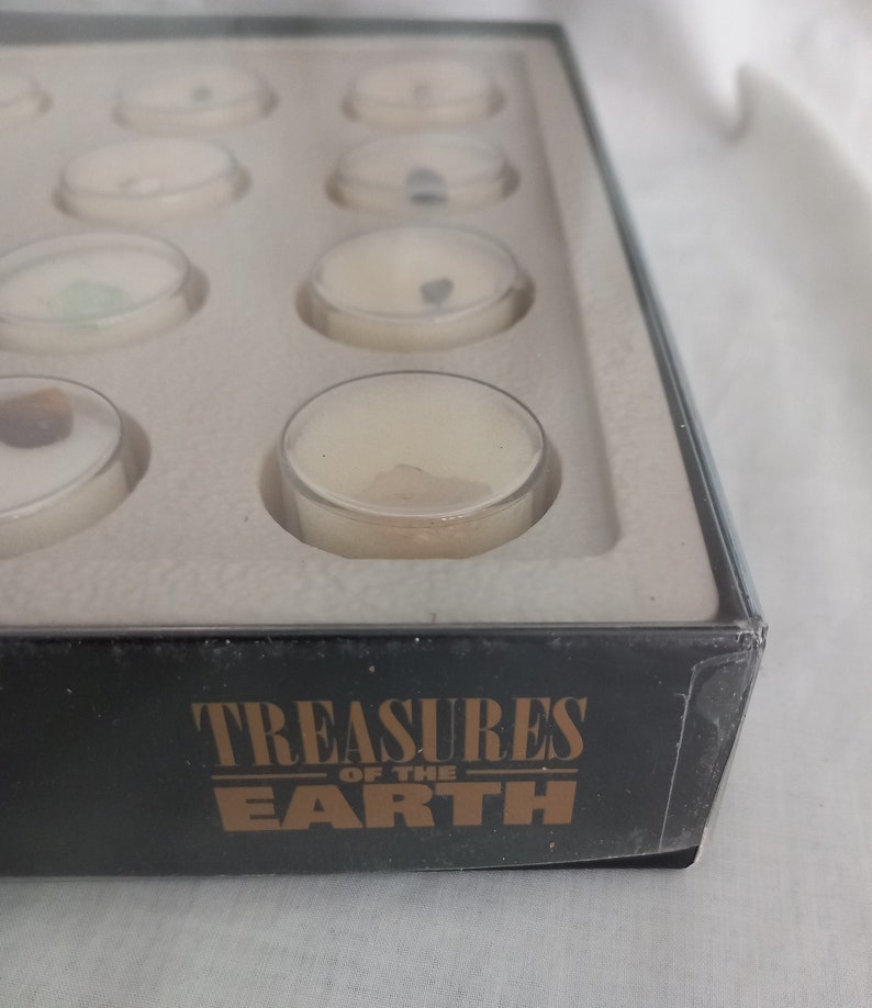Puede incluir: Una caja negra con la inscripci&oacute;n dorada "Treasures of the Earth", que contiene peque&ntilde;os recipientes con tapa transparente. Cada recipiente contiene una muestra de tesoros de la tierra. Los recipientes est&aacute;n dispuestos en filas dentro de un inserto blanco.