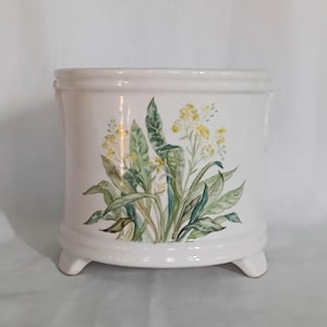 Puede incluir: Una maceta de cerámica blanca con un diseño floral de flores amarillas y hojas verdes. La maceta tiene forma cuadrada con esquinas redondeadas y se apoya en tres pequeños pies. El diseño floral está pintado en la parte delantera.