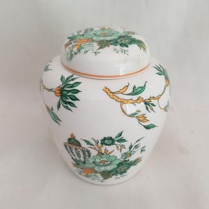 May include: A white ceramic jar with a lid, decorated with green and orange floral patterns. The jar has a rounded shape and is decorated with detailed botanical designs, including leaves and flowers. The lid has a matching floral pattern and an orange trim.