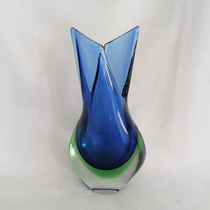 May include: A striking glass vase with a unique form. The vase showcases a bold colour palette, with a dark blue upper part and a green base. The design incorporates two upward-curving, pointed elements at the top.