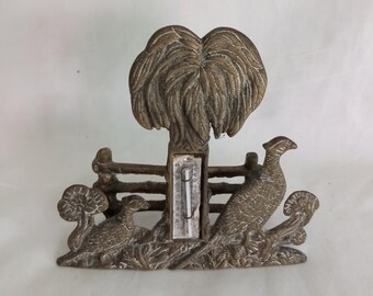 May include: Brass thermometer with a pheasant and a tree design. The thermometer is framed by a fence and has a white background.