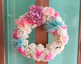 May include: A white and pink floral wreath with light blue accents hangs on a wooden door. The wreath is made of artificial flowers and is a great addition to any home decor.