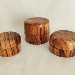 Puede incluir: Tres recipientes redondos de madera con tapa, cada uno con un patrón de veta natural distintivo en tonos marrón claro y oscuro. Los recipientes están dispuestos en grupo, mostrando un acabado liso y pulido en las superficies exteriores.