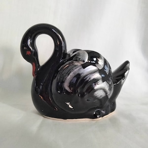 Puede incluir: Una figura de cisne de cerámica negra con cuello curvo y acento rojo en el pico. El cuerpo del cisne tiene pinceladas plateadas y blancas. La figura está sobre una superficie blanca.