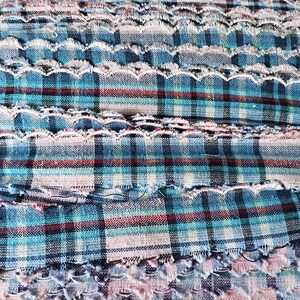 Puede incluir: Primer plano de una tela texturizada con un patrón de cuadros en tonos azules, rojos y blancos. La tela tiene un borde festoneado y una textura deshilachada, lo que sugiere una estética artesanal o vintage. La tela es adecuada para ropa o decoración del hogar.