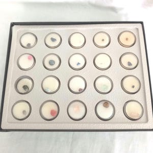 Puede incluir: Una bandeja rectangular negra con 20 peque&ntilde;os recipientes redondos y transparentes. Cada recipiente contiene una peque&ntilde;a gema de colores colocada en cera blanca. Las gemas son de varios colores, incluyendo rosa, azul, verde y marr&oacute;n.